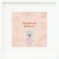 An inspirational print with a graphic of Claire the koala on a blush-colored background decorated with leaves and the words “Daydream believer” in orange-pink.