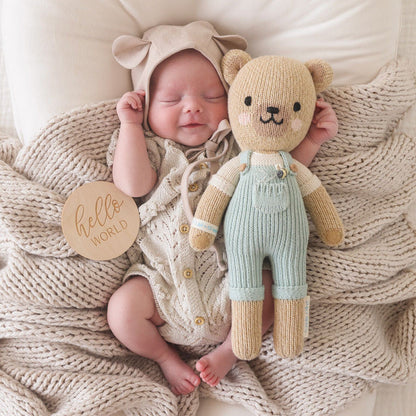 A sleeping baby in a bear-eared bonnet lying on a knit blanket next to Charlie the honey bear. Beside the baby is a small wooden sign with text that says "hello world"