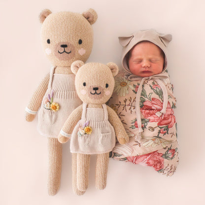 A sleeping baby, wrapped in a floral swaddle, lying next to two Golide the honey bears, in regular and little sizes.