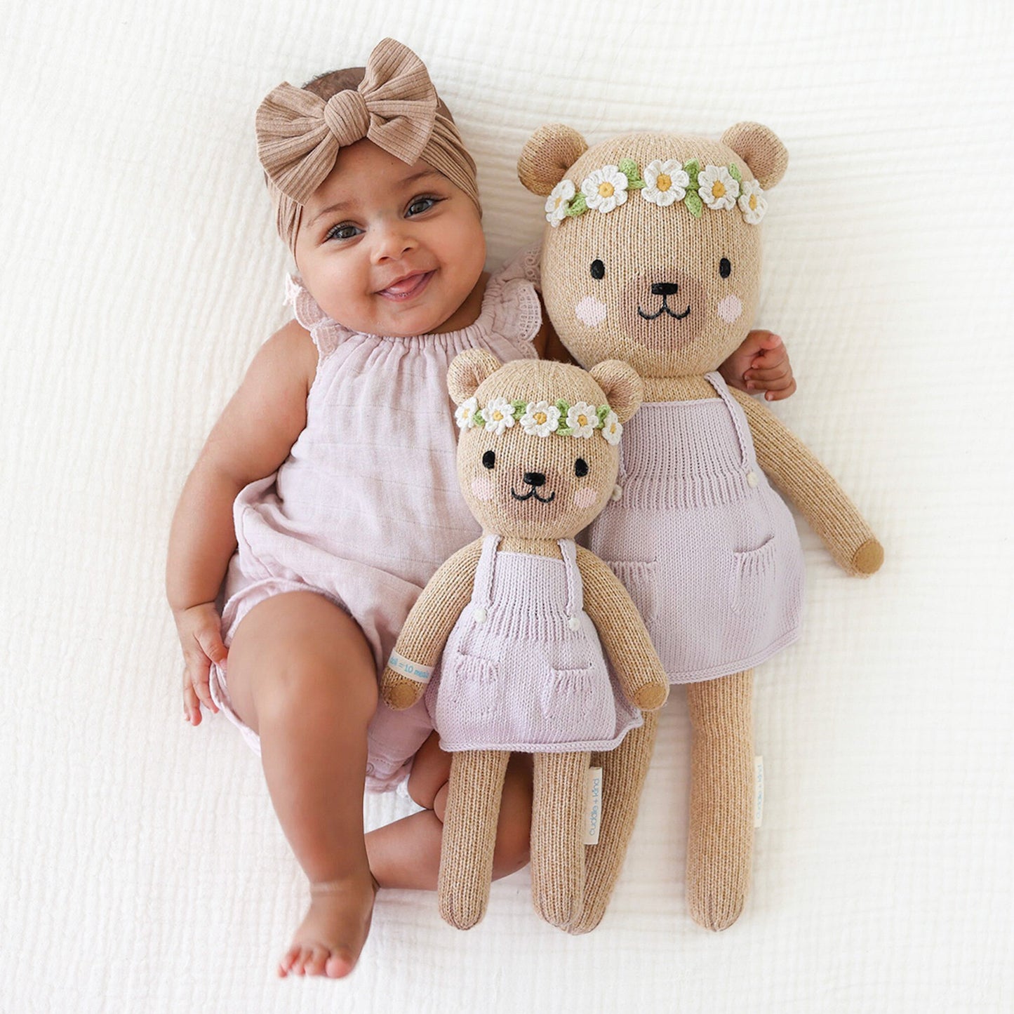 A smiling baby in a lavender onesie with their arms around two Olivia the honey bear stuffed dolls, in the little and regular sizes.