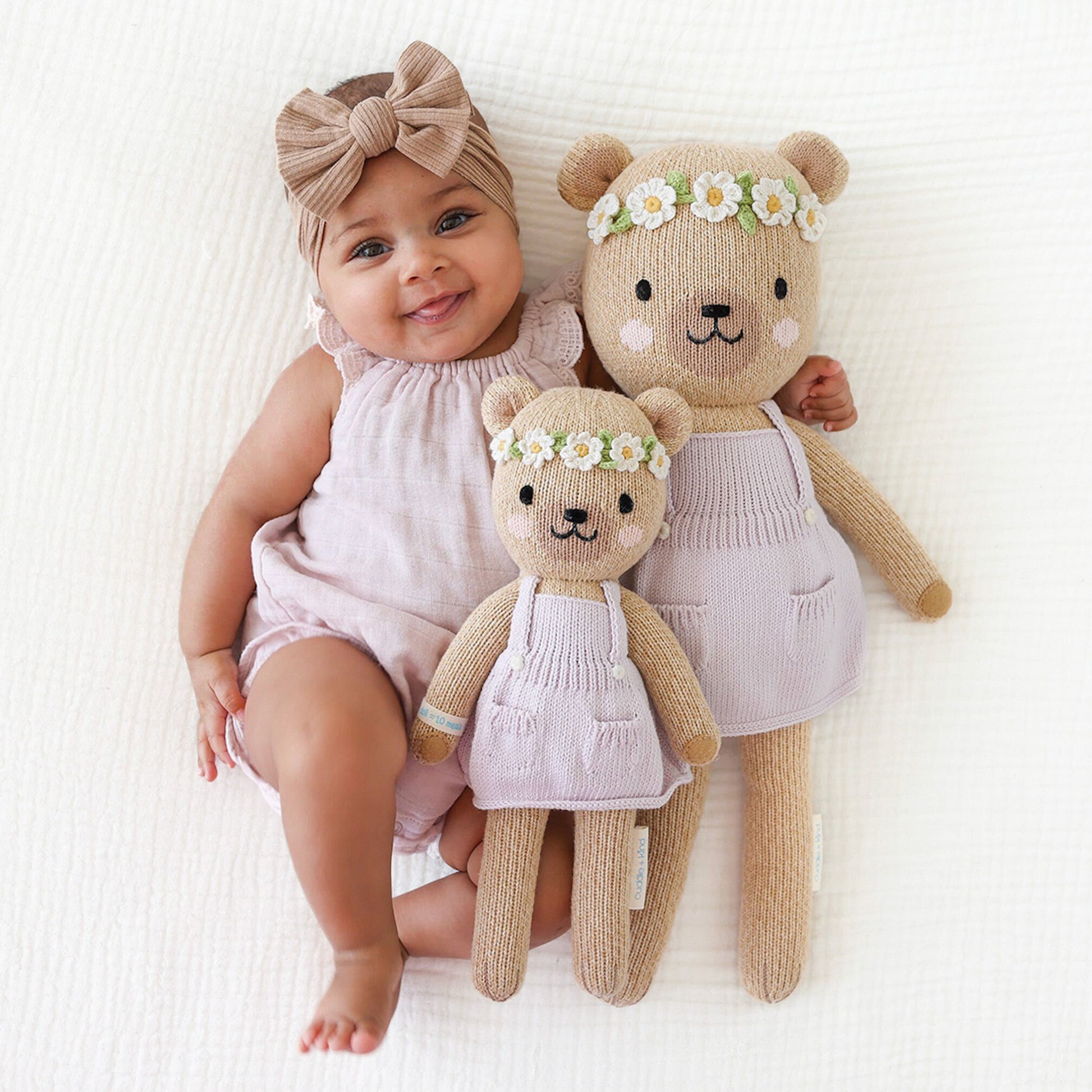 A smiling baby in a lavender onesie with their arms around two Olivia the honey bear stuffed dolls, in the little and regular sizes.