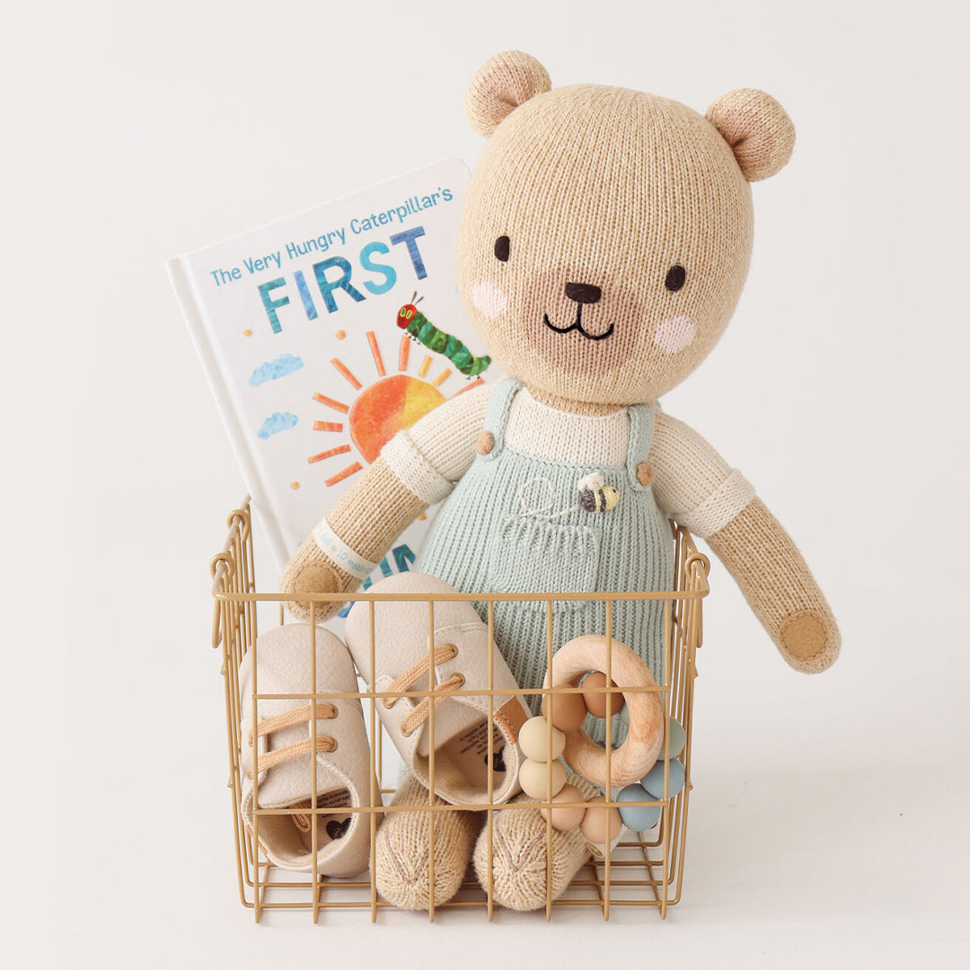 Charlie the honey bear sitting in a wire basket, alongside a children’s book, a pair of booties and a wooden infant’s toy.