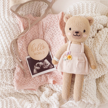 Goldie the honey bear lying on a hand-knit blanket, next to an ultrasound photo, a knit romper and a small wooden sign with text that says "hello world"