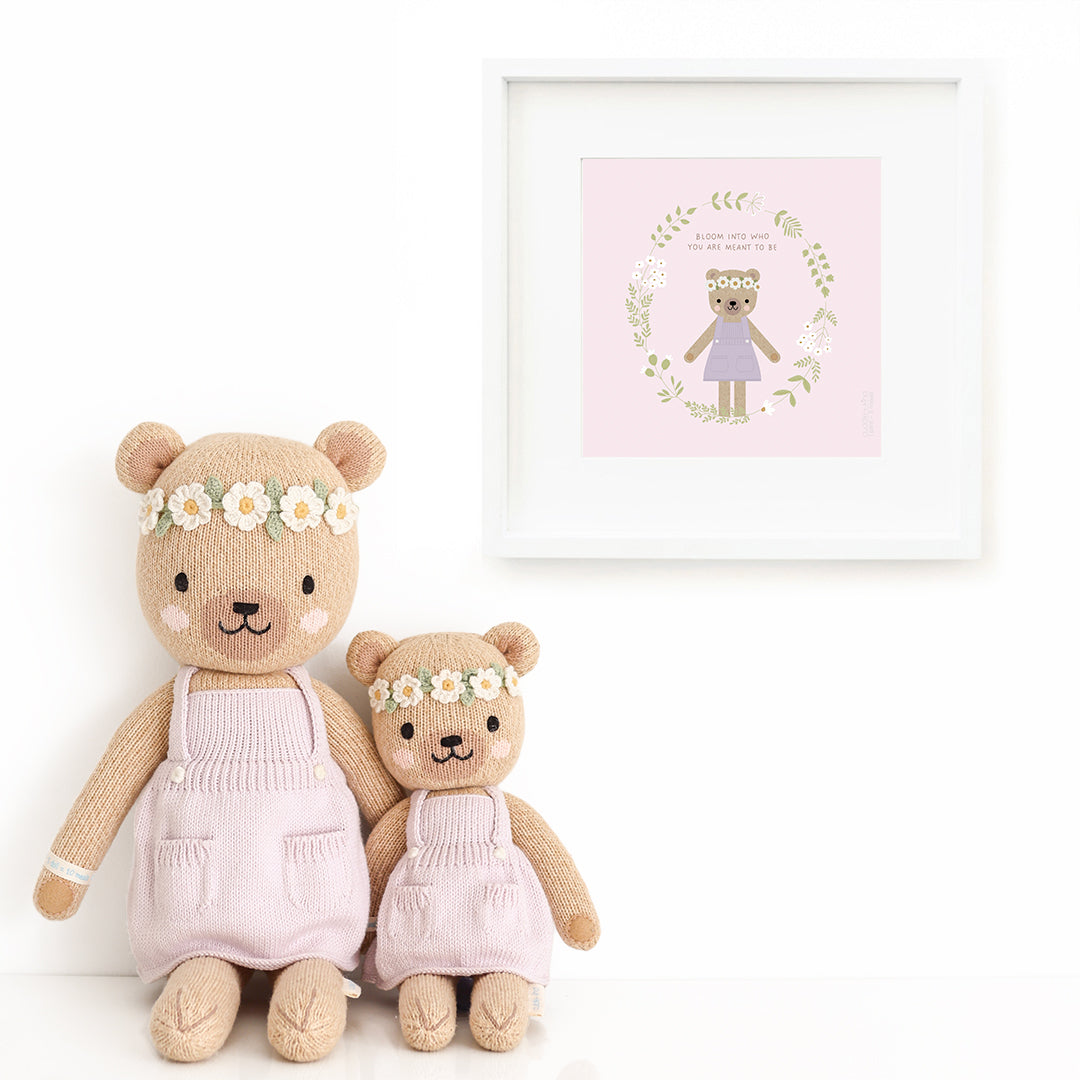 Two Olivia the honey bear stuffed dolls sitting beside a framed print with a picture of Olivia, with text that says "Bloom into who you are meant to be."