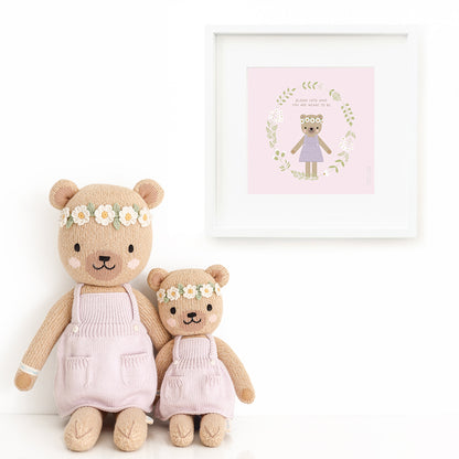 Two Olivia the honey bear stuffed dolls sitting beside a framed print with a picture of Olivia, with text that says "Bloom into who you are meant to be."