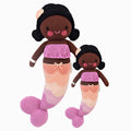 Maya the mermaid in the regular and little sizes, shown from the front. Maya has black hair with a pink shell on her head. She has a tasselled purple top, and the scales on her tail are purple, pink, peach and cream.