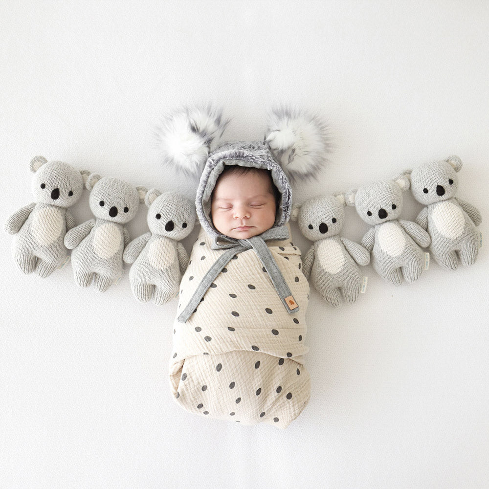 A sleeping baby in a koala hood with six baby koala stuffed dolls.