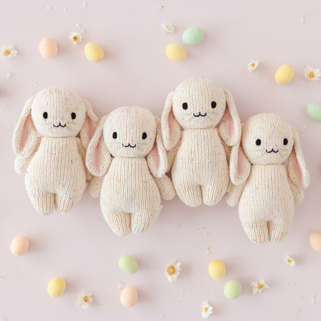 A group of four baby bunny in confetti stuffed dolls surrounded by daisies and pastel Easter eggs.