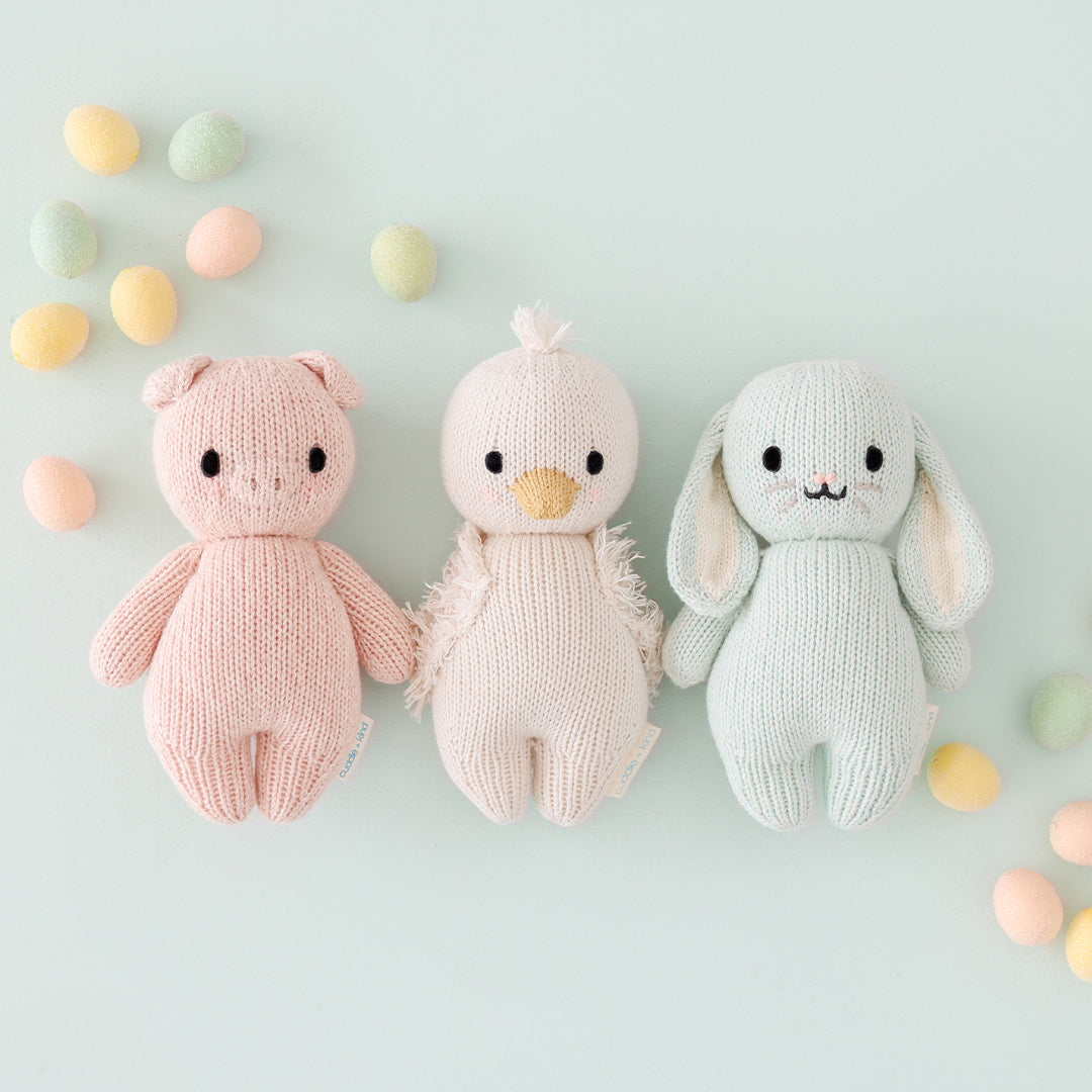 A group of three baby animals, baby piglet, baby gosling and baby bunny in mint, lying alongside pastel Easter eggs.