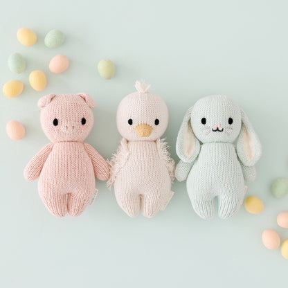 A group of three baby animals, baby piglet, baby gosling and baby bunny in mint, lying alongside pastel Easter eggs.