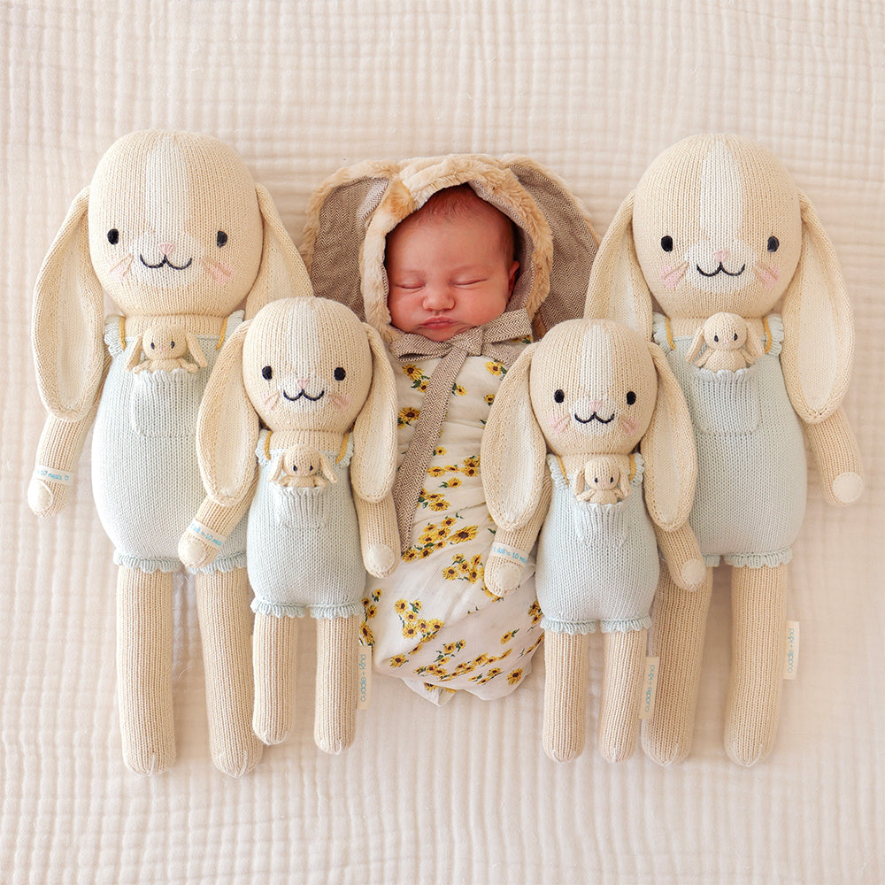 A sleeping baby in a bunny eared-bonnet lying side-by-side with four Briar the bunny dolls in the regular and little sizes.