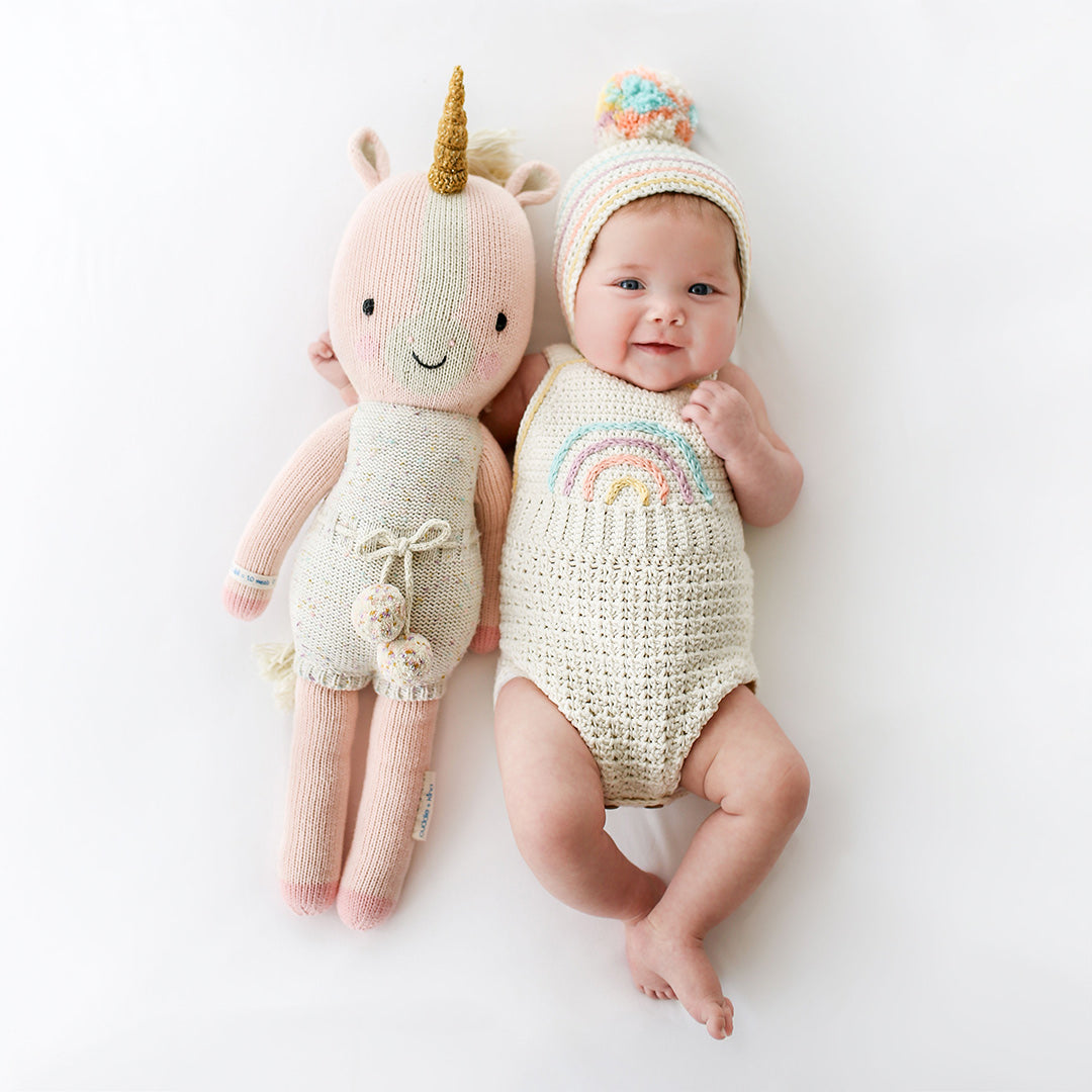 A smiling baby in a knit hat and romper with their arm around an Ella the unicorn stuffed doll.