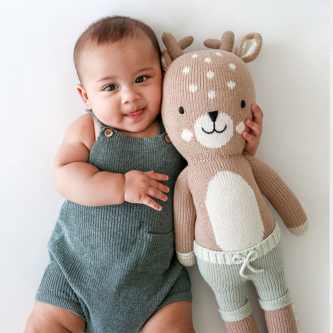 A smiling baby with his arm around an Elliott the fawn doll.