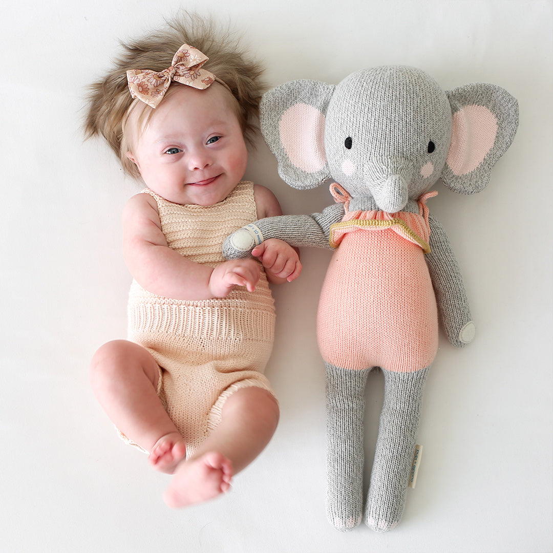 A smiling baby, wearing a sparkly hair bow, holding hands with Eloise the elephant.