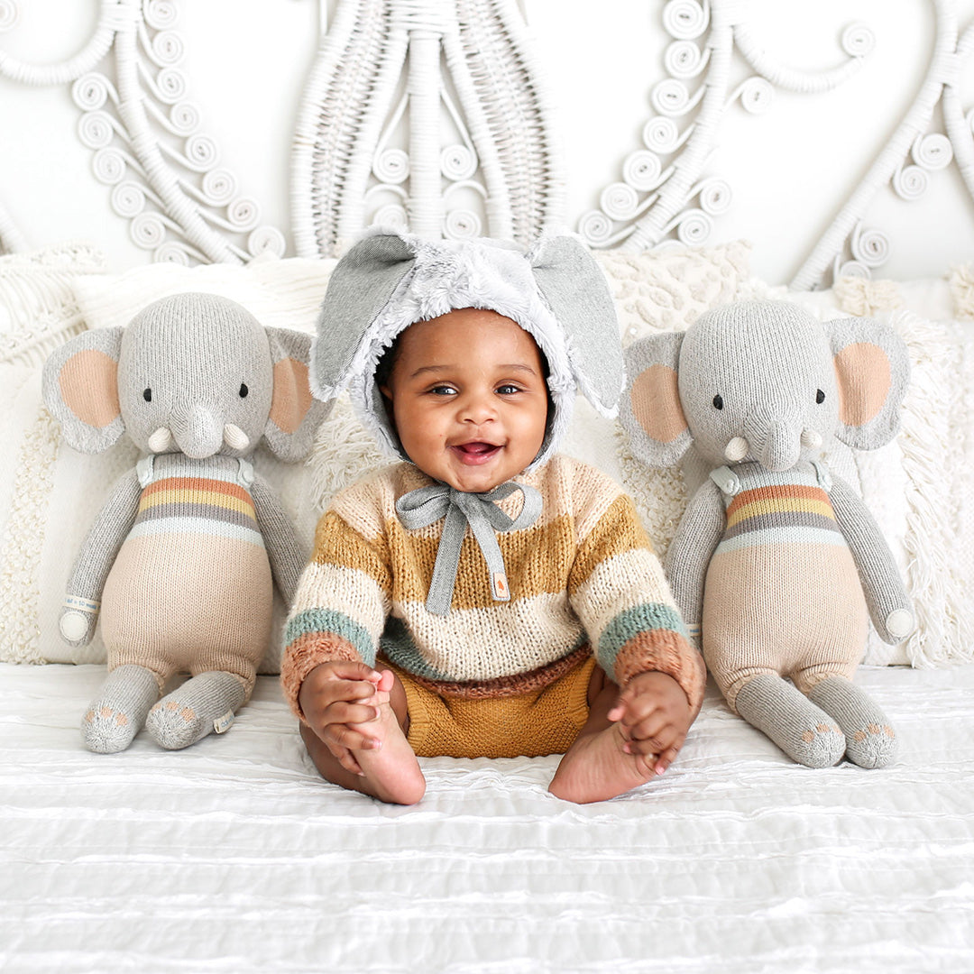 A baby wearing an elephant hood sitting on a bed next to two Evan the elephant stuffed dolls.