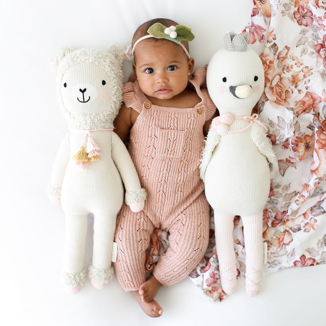 A baby with their arms around two cuddle and kind stuffed dolls, Lola the llama and Harlow the swan.