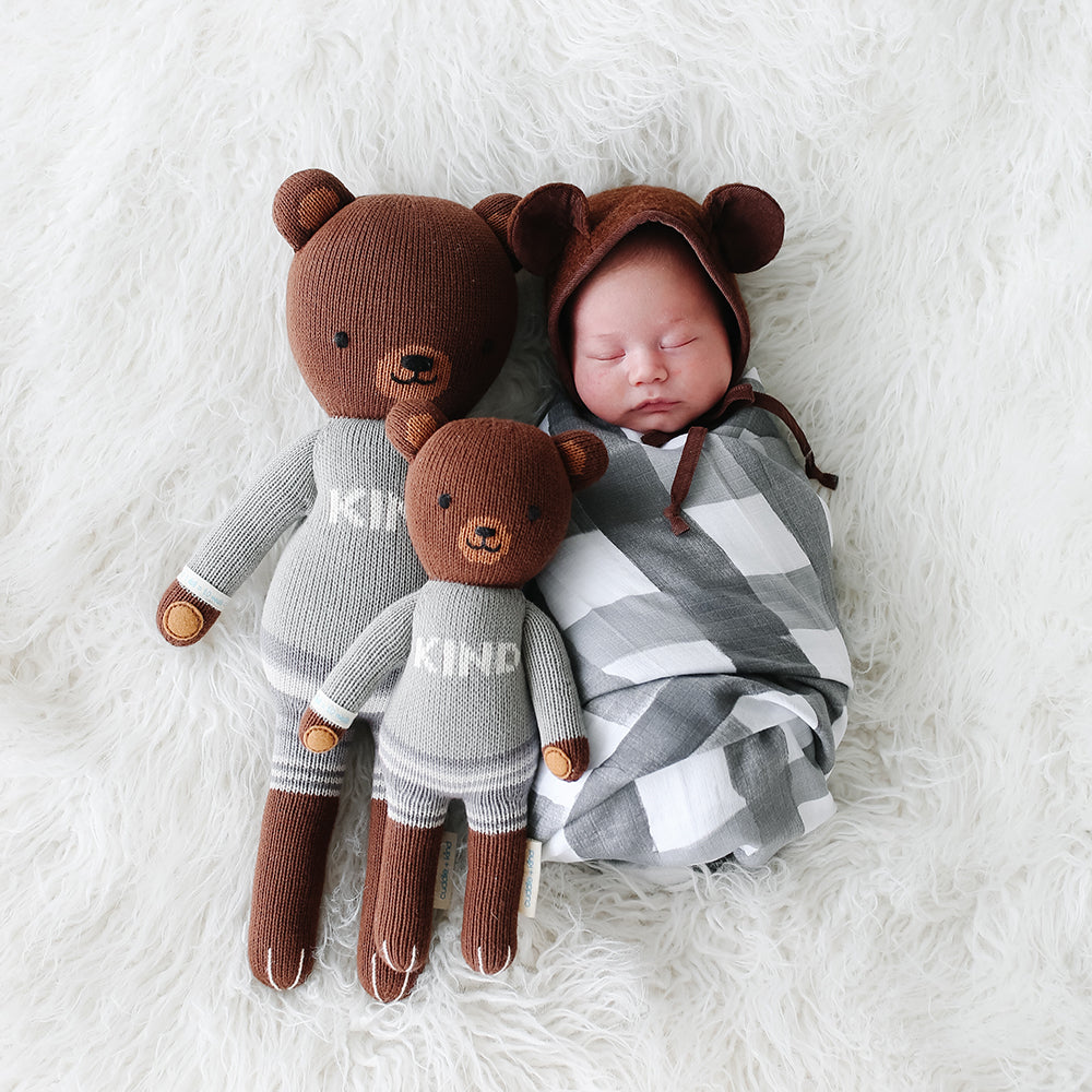 A sleeping baby in a bear hood snuggled up with two Oliver the bear dolls in the regular and little sizes.