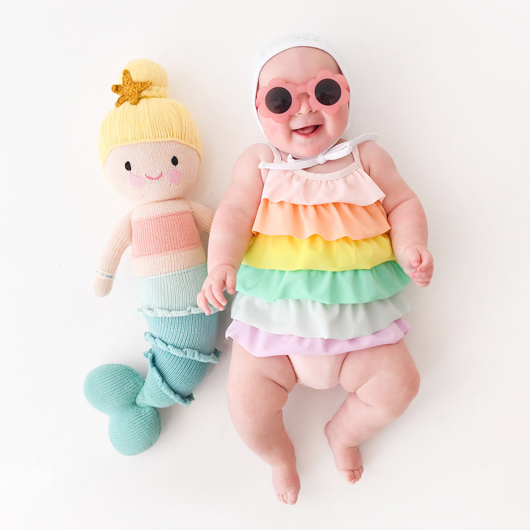 A laughing baby wearing a multi-coloured ruffled swimsuit and floral sunglasses with a Skye the mermaid stuffed doll.