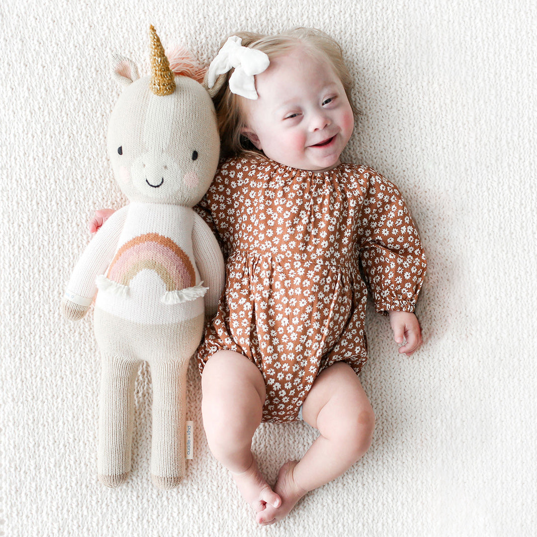 A baby wearing a floral romper with her arm around a Zara the unicorn stuffed doll.