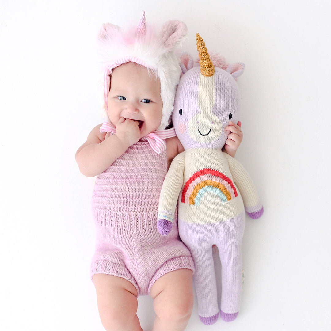 A baby in a unicorn hat with a Zoe the unicorn doll in the regular size.