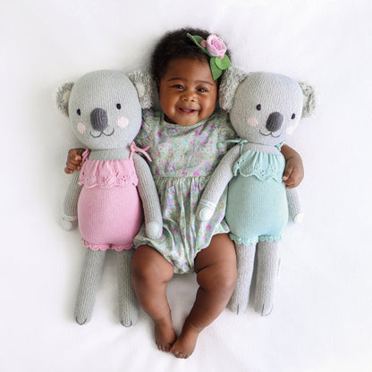 A smiling baby with two Claire the koala stuffed animals in pink and mint.