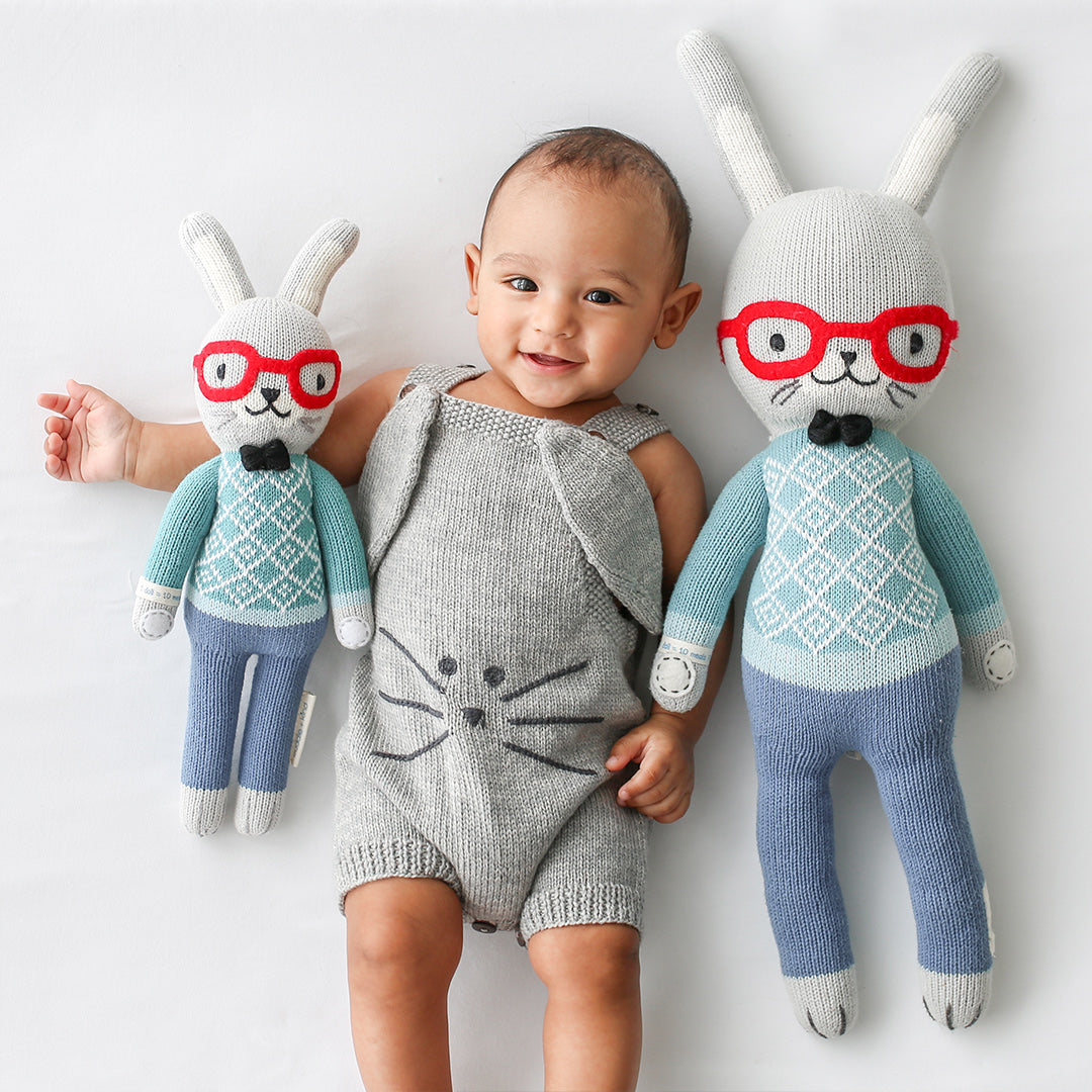 A smiling baby lying arm-in-arm with two Benedict the bunny dolls in the regular and little sizes.