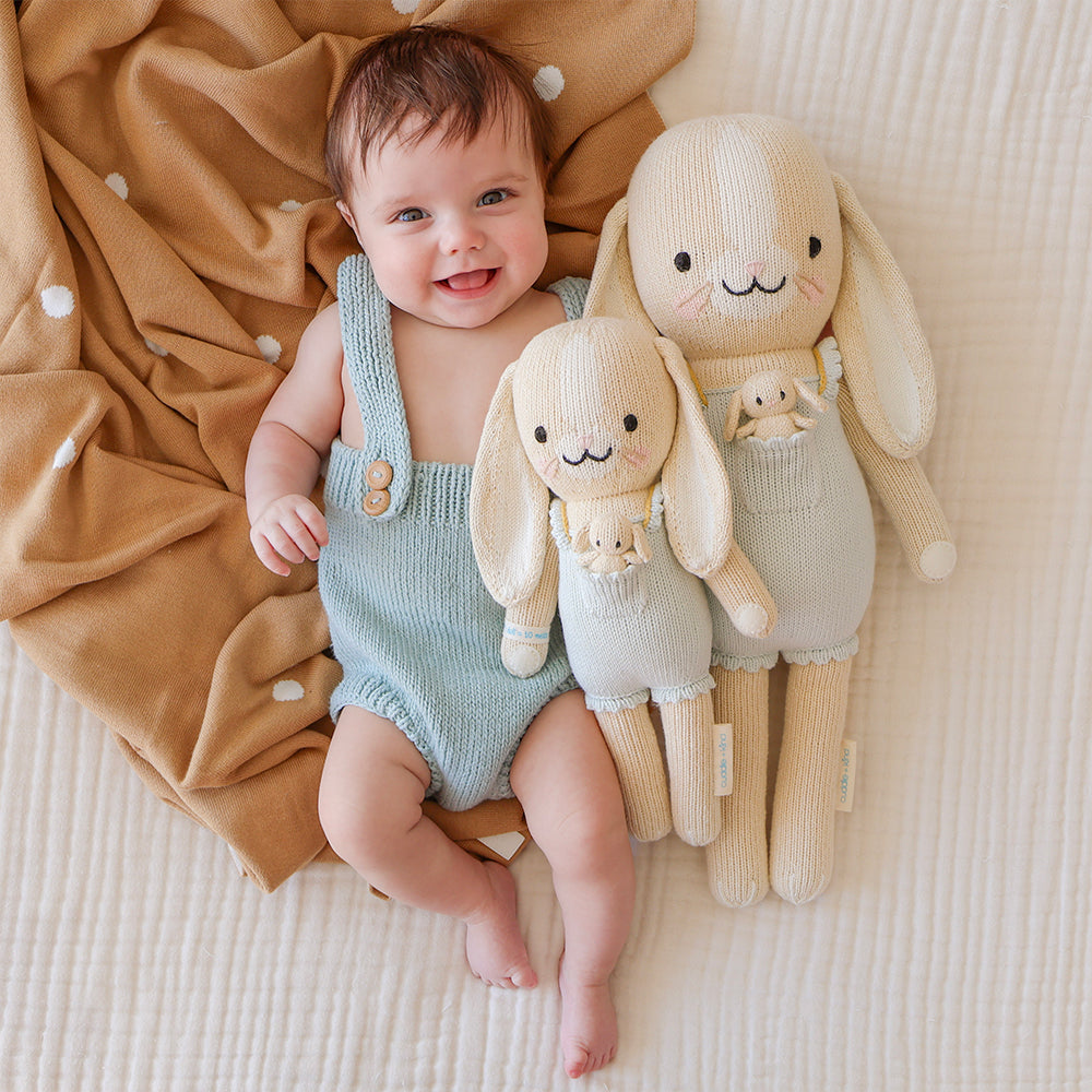 A smiling baby snuggled beside two Briar the bunny dolls in the regular and little sizes. The baby is wearing a knit overall romper, similar to Briar the bunny’s romper.