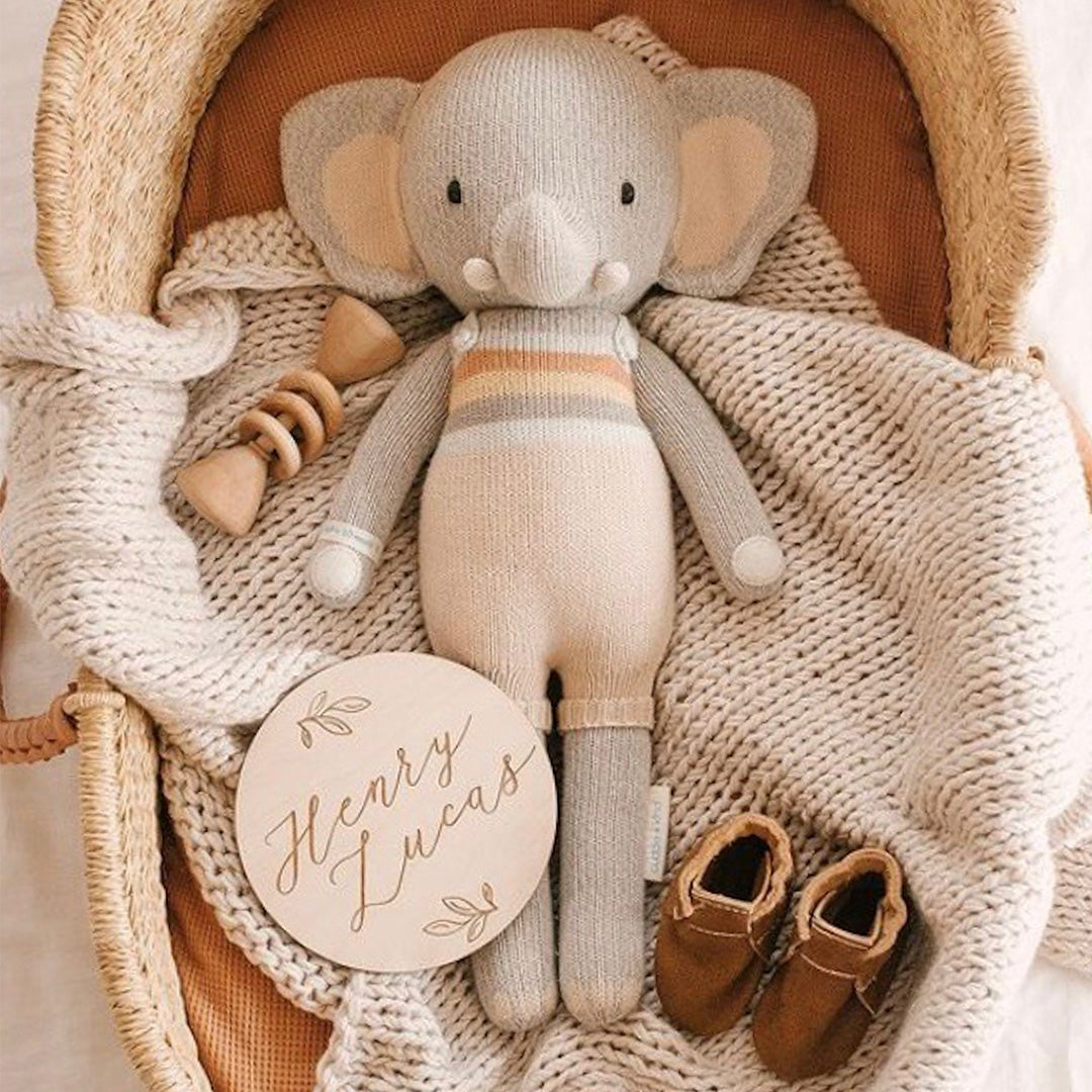 Evan the elephant laying in a wicker baby basket, alongside a pair of infant's booties, a wooden rattle and a wooden sign with text on it that says "Henry Lucas".