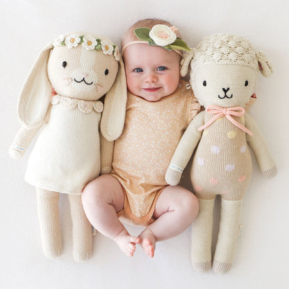 A smiling baby with their arms around two cuddle and kind stuffed dolls, Hannah the bunny and Lucy the lamb.