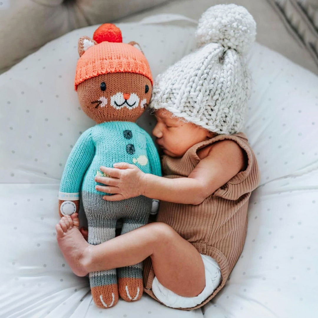 A sleeping baby snuggled up with their arms and legs wrapped around Maximus the cat. The baby is wearing a pom-pom hat similar to Maximus’.