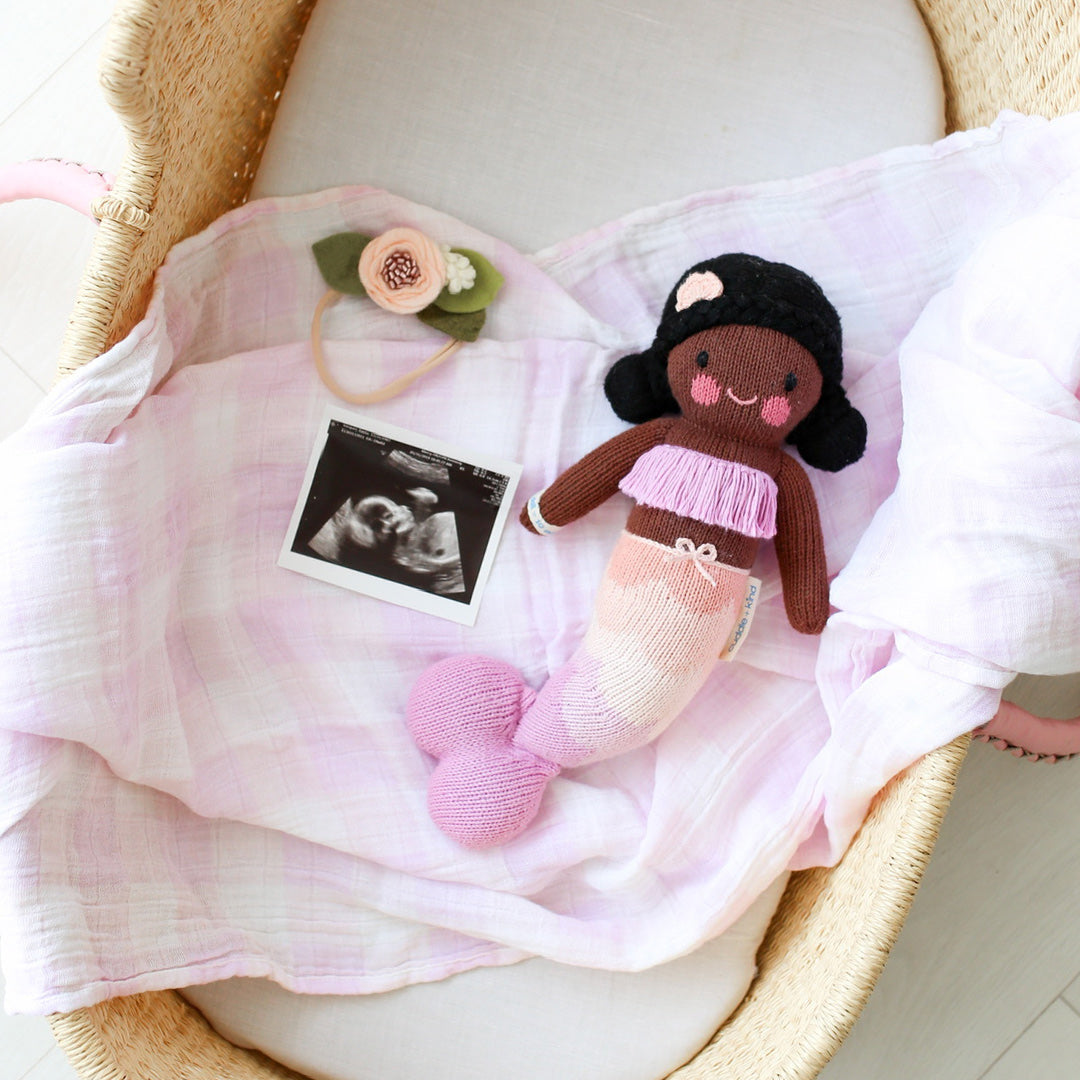 Maya the mermaid lying on a baby blanket inside a bassinet. Beside her are a baby's floral headband and an ultrasound image.