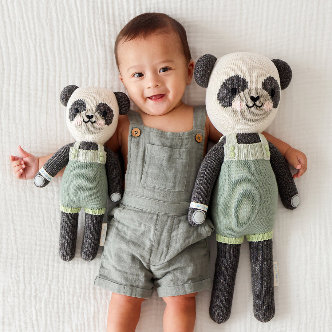 A smiling baby, wearing a green romper, with their arms around two Paxton the panda stuffed dolls, in the regular and little size.