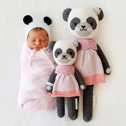 A swaddled, sleeping baby, lying beside two Polly the panda stuffed dolls, in the regular and little sizes.