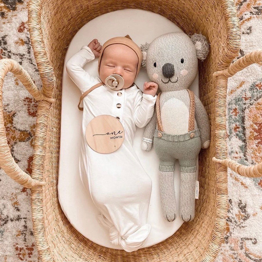 A sleeping baby, stretched out in a bassinet beside Quinn the koala. A little wooden sign resting on the baby’s tummy has text that says “one month.”