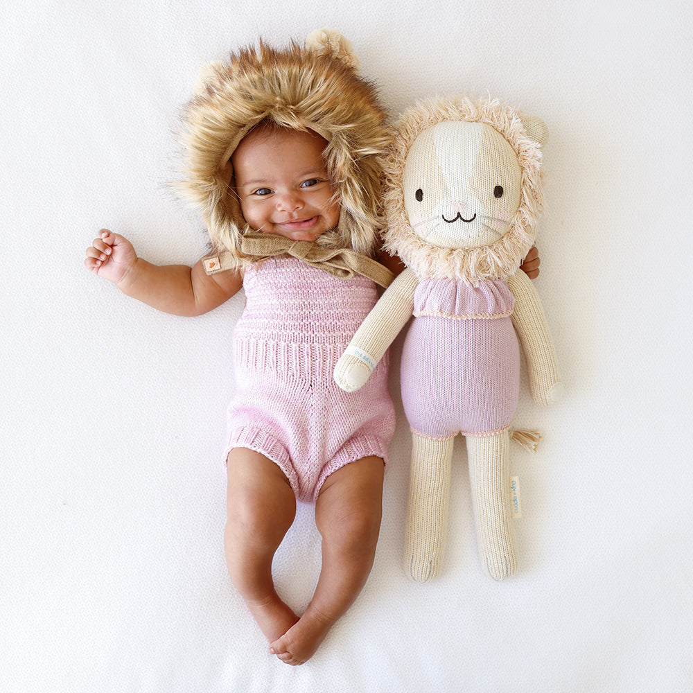 A smiling baby in a fluffy lion hood with her arm around a Savannah the lion doll. The baby is wearing a purple knitted romper to match Savannah.