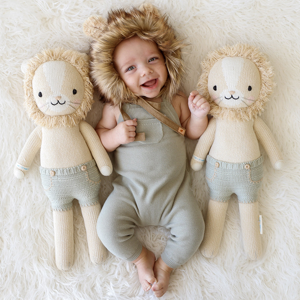 A laughing baby in a fluffy lion hood with two Sawyer the lion stuffed dolls.