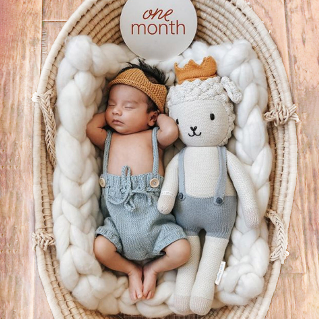 A sleeping baby with a Sebastian the lamb doll in the regular size. The baby has a knitted crown and an outfit to match Sebastian.