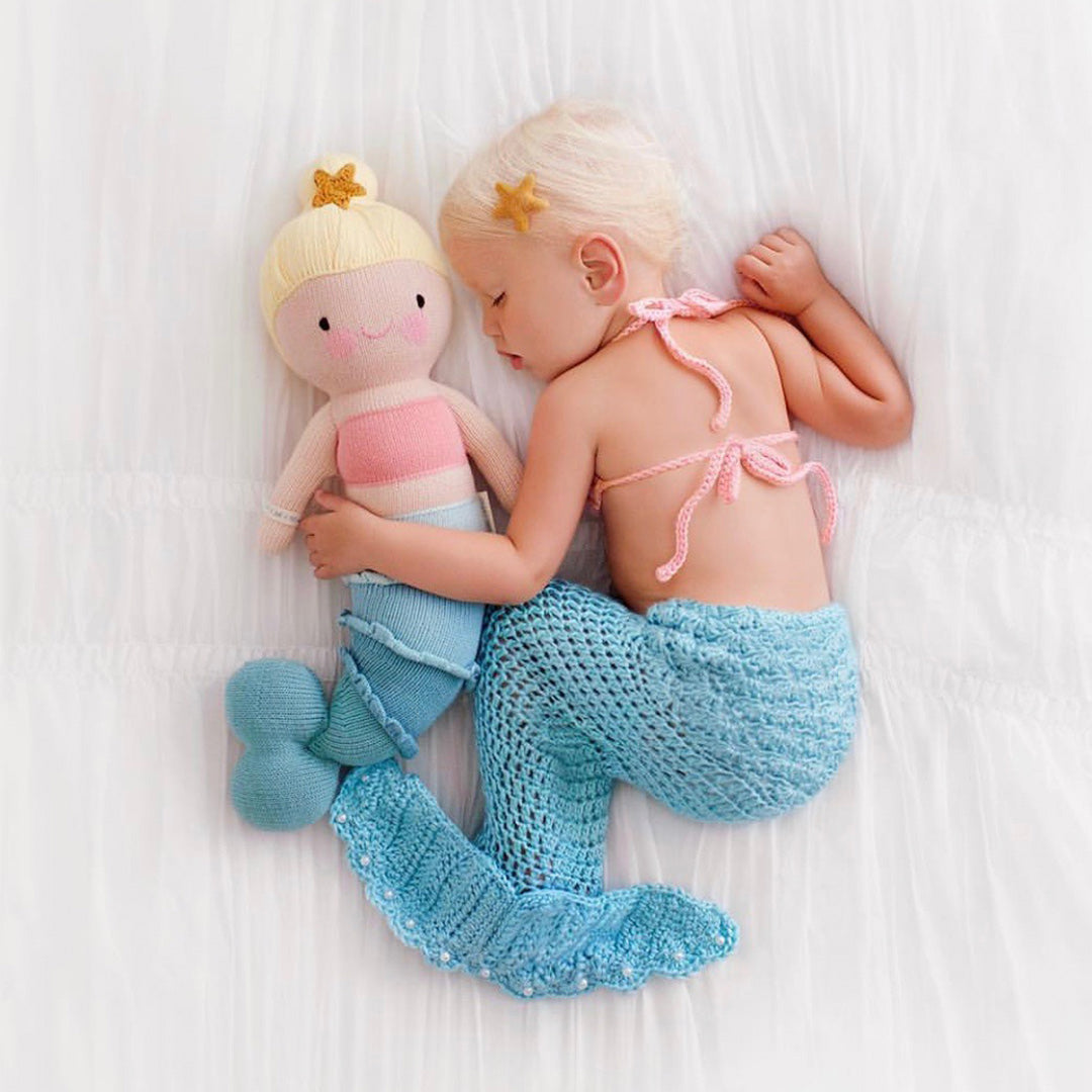 A sleeping child with her arm around Skye the mermaid. The child is wearing a knit mermaid tail and pink top that matches Skye the mermaid.