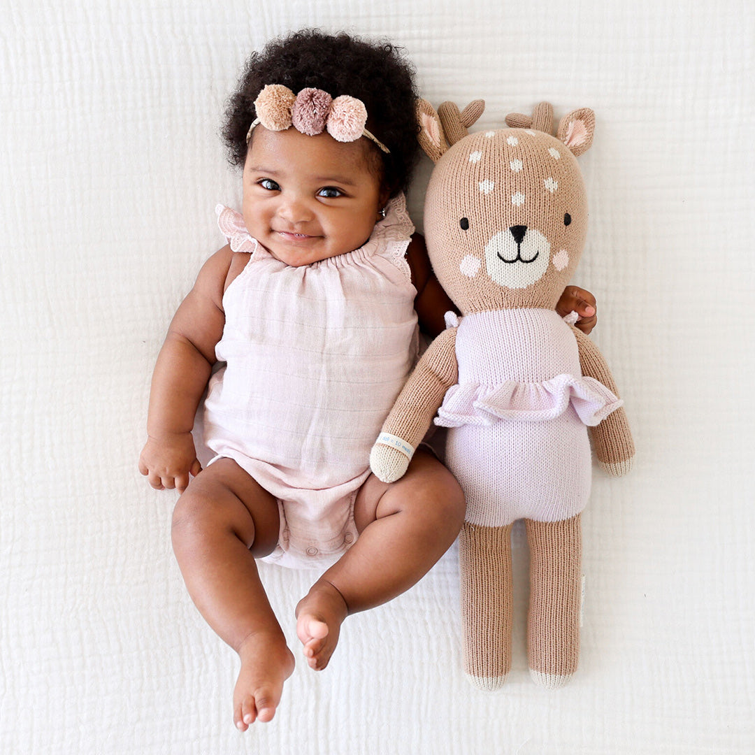 A smiling baby with her arm around Violet the fawn. The baby is wearing a purple onesie to match Violet.