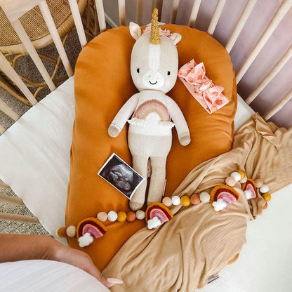 Zara the unicorn lying in a crib alongside a decorative felt garland and an ultrasound image. A woman holding her baby bump can be seen above the crib.