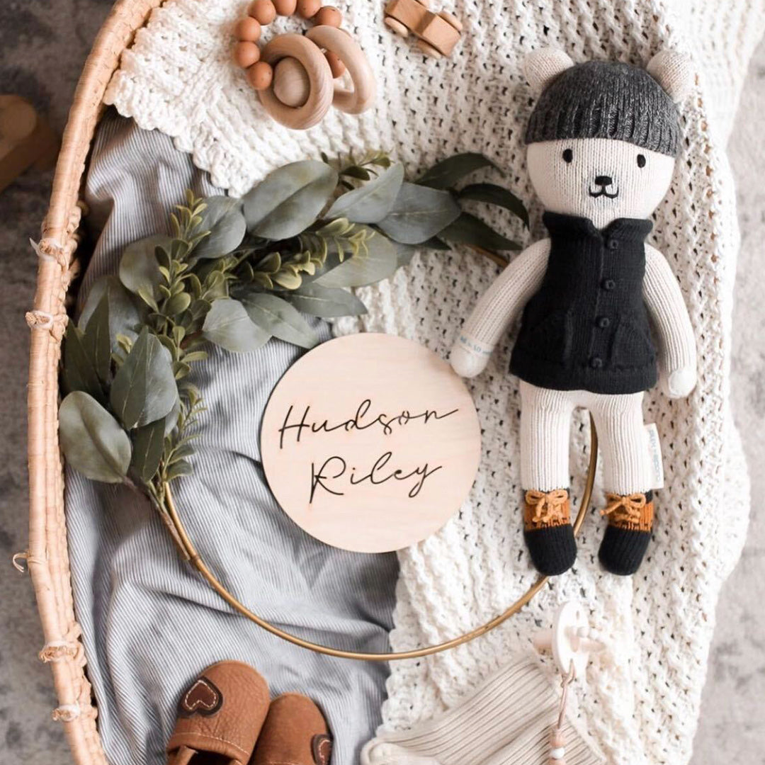 Hudson the polar bear lying in a wicker bassinet alongside a wooden sign displaying the name Hudson Riley. There are baby toys and booties in the bassinet, along with a decorative wreath.