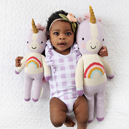 A baby with her arms around two Zoe the unicorn dolls in the little and regular sizes.