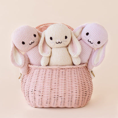 Three baby animal stuffed dolls peeking out of a pink basket: baby bunny in oatmeal, lilac and rose.