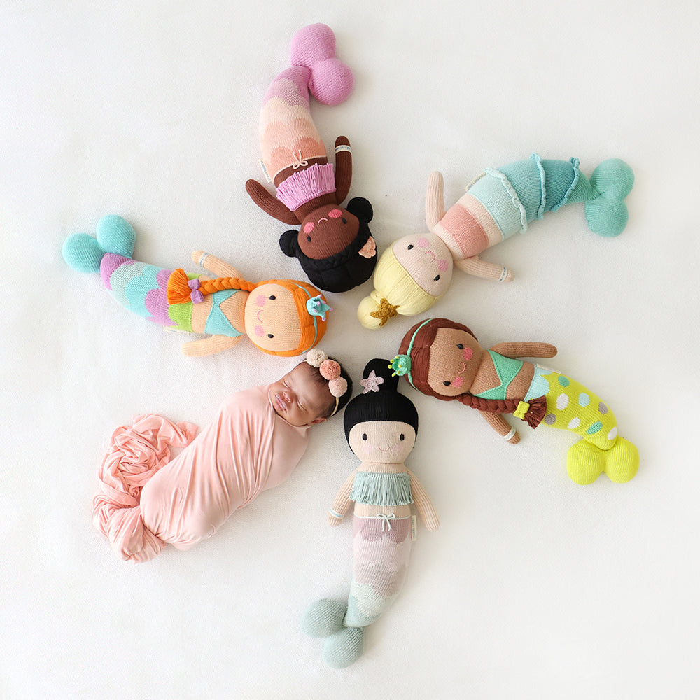 A sleeping baby lying in a circle head-to-head with five mermaid dolls: Isla, Maya, Skye, Pearl and Luna.