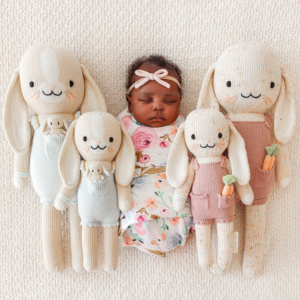 A sleeping baby lying in between four dolls, Briar the bunny and Harper the bunny, in the little and regular sizes.