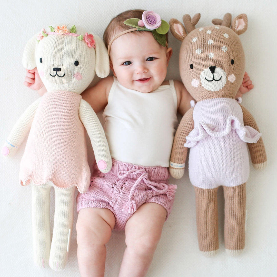 A smiling baby wearing a floral headband with their arms around a Charlotte the dog stuffed doll and a Violet the fawn stuffed doll.