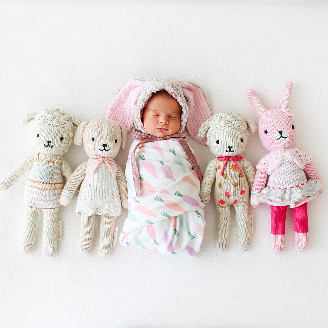 A baby in a bunny bunny-eared bonnet lying between four cuddle and kind stuffed dolls.