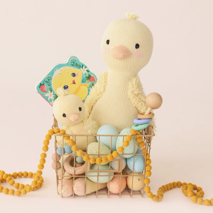 A Finley the duckling stuffed doll and a baby duckling stuffed animal in a wire basket, along with pastel eggs, a children's book and a few wooden toys and ornaments.