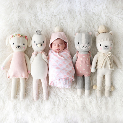 A sleeping baby, wearing a pom pom bonnet, nestled between four cuddle and kind stuffed dolls.