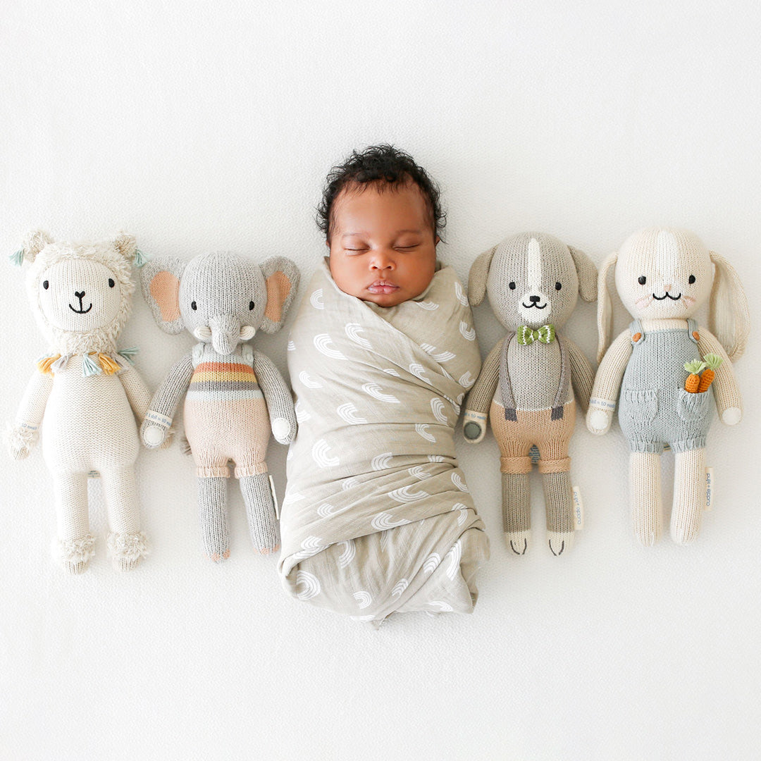 A sleeping baby, wrapped in a swaddle, surrounded by four cuddle+kind stuffed dolls.